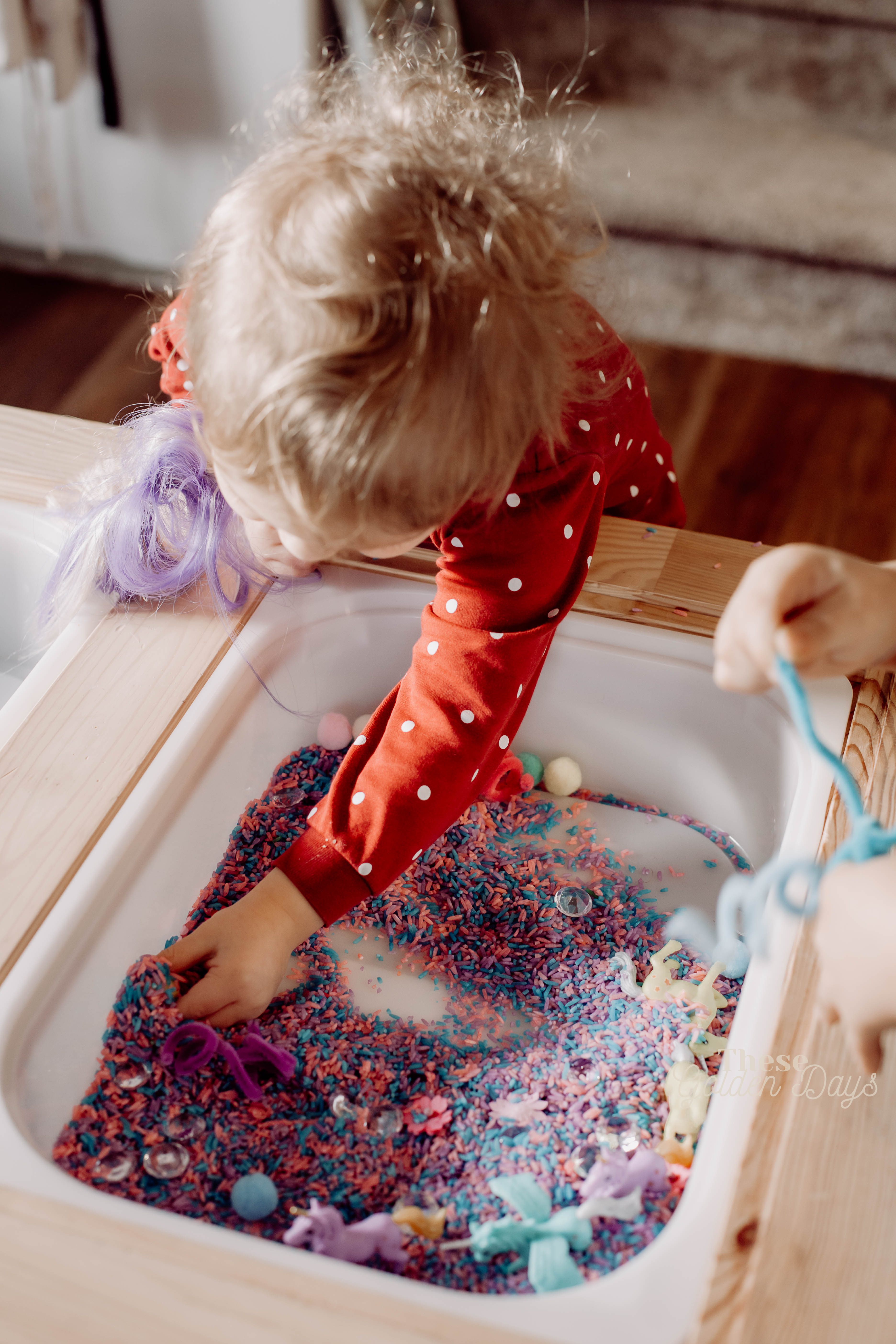 Child playing with coloured rice, unicorns, pipe cleaners, jewels and pom poms in a sensory table.