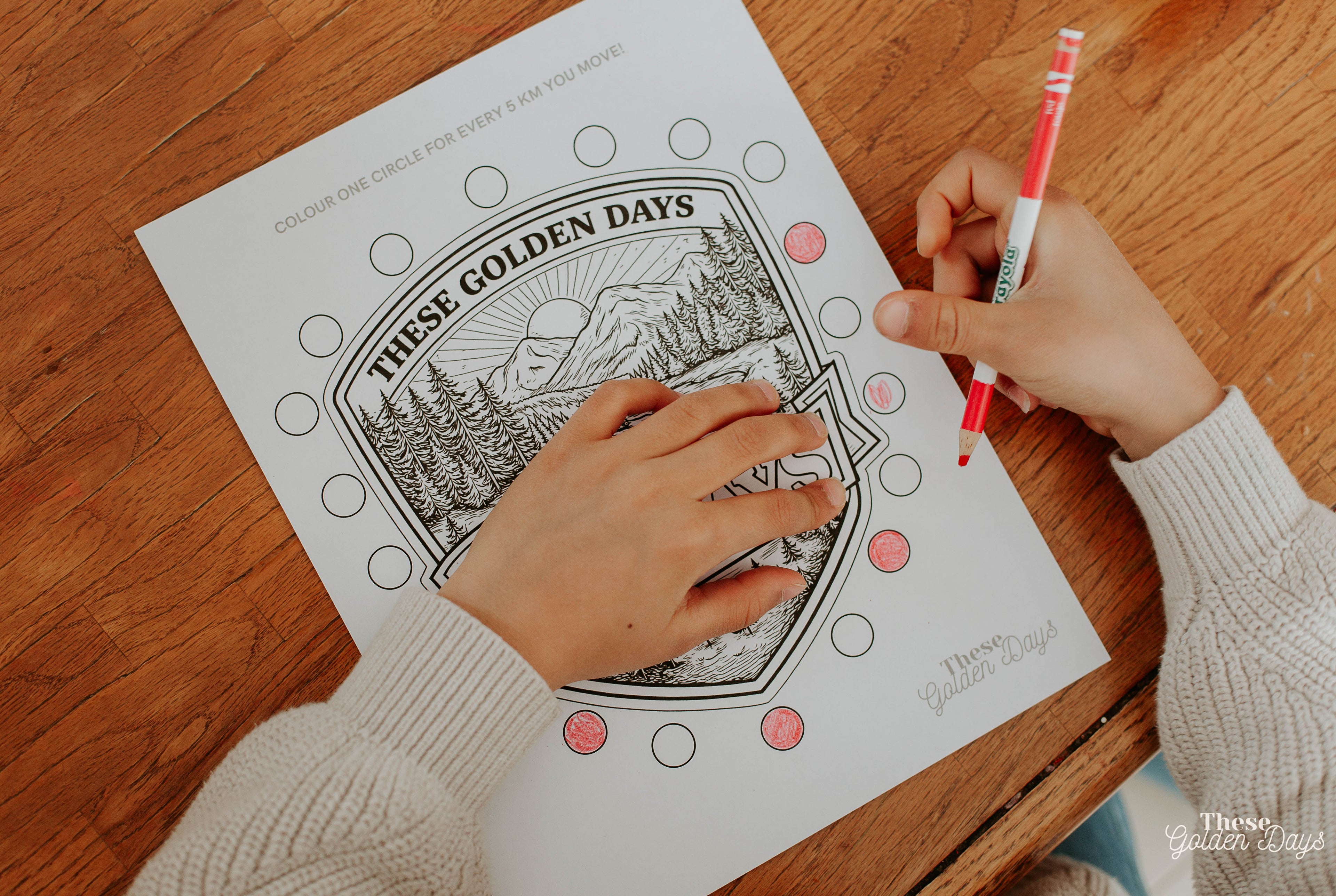 Person coloring a 'These Golden Days' coloring book with a red marker on a wooden table.
