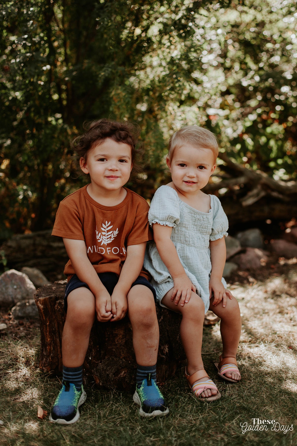 Two children sitting on a log in a natural setting with greenery.