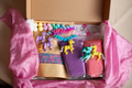 Open box with toy unicorns, pom poms, jewels, pip cleaners, two packs of playdough and a pack of rainbow rice.  bow is sitting on a pink silk. 