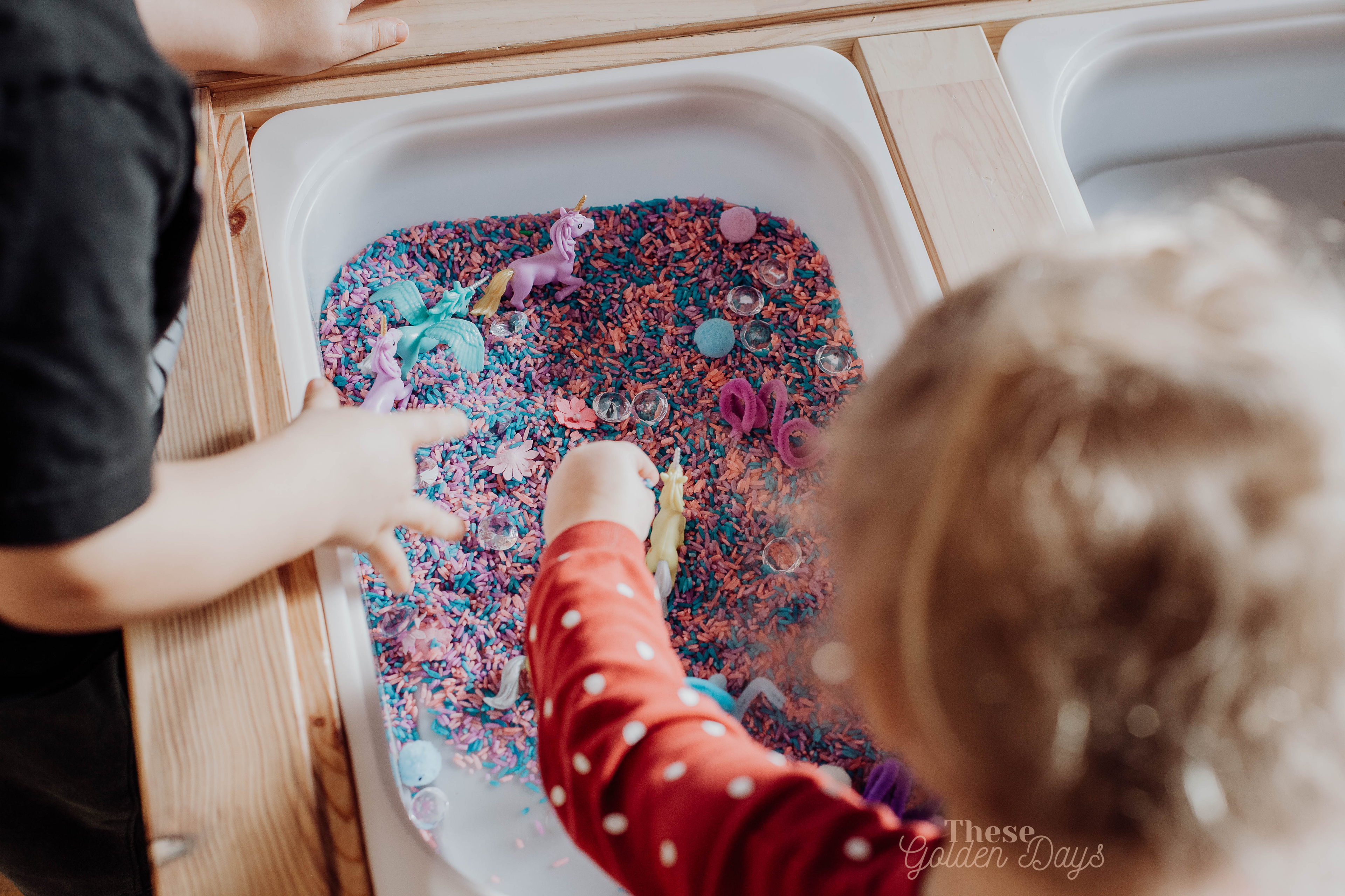 Children using a sensory table to play with rainbow rice, toy unicorns, pom poms, jewels and pipe cleaners. 